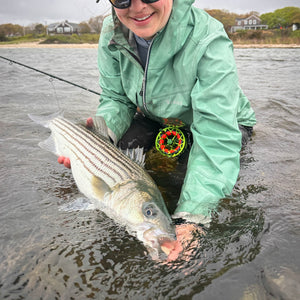 Angler kneeling in shallow coastal water while holding a striped bass, with a colorful fly reel clipped to their jacket.