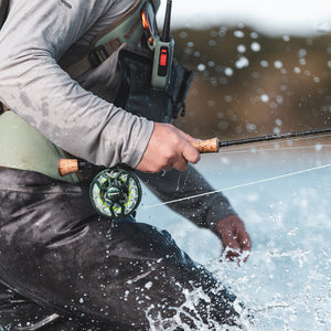 Angler wearing waders fly fishing in shallow water, holding a fly rod with a reel as water splashes around their legs.