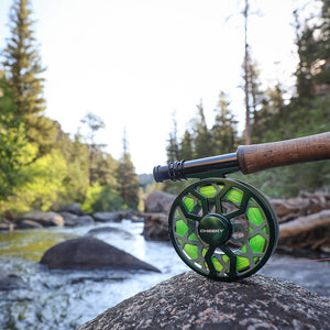 Fly fishing reel with green line resting on a rock beside a flowing river.