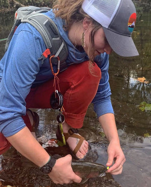 Angler kneeling in a creek, adjusting a fly on a trout’s mouth while wearing waders, sandals, and a trucker hat.