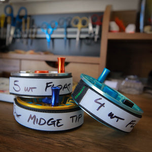 Three fly reels labeled with 5-wt float, 5-wt midge tip, and 4-wt float on a workbench with fly-tying tools in the background.