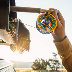 Person loading a fly rod with a yellow-and-blue fly reel into a rooftop rod carrier at sunrise.