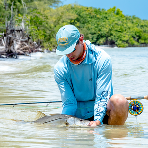 Angler kneeling in shallow tropical water holding a large fish while a fly rod with a blue-and-gold reel rests nearby.