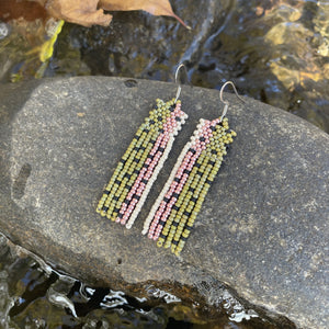 A pair of long beaded earrings in green, pink, and cream tones resting on a wet rock beside a gently flowing stream.