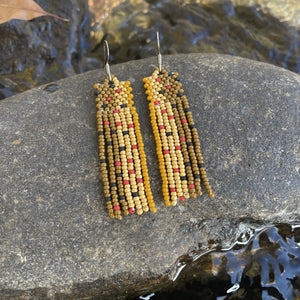 Pair of handmade beaded earrings in gold, red, and brown tones displayed on a rock next to flowing creek water.
