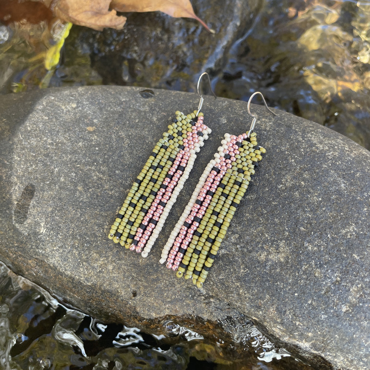 A pair of long beaded earrings in green, pink, and cream tones resting on a wet rock beside a gently flowing stream.