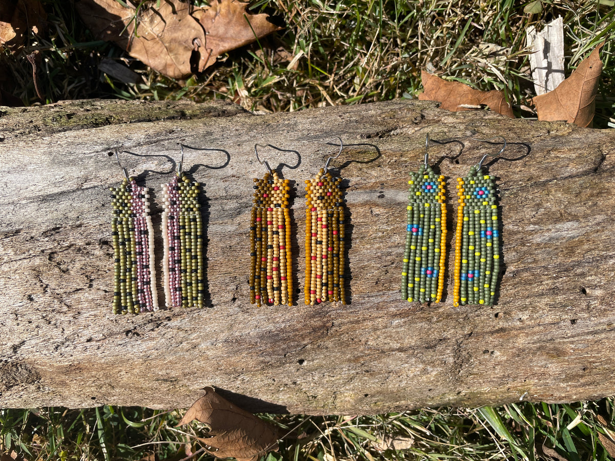 Three pairs of long, beaded earrings in different color patterns arranged on a sunlit log in grass.