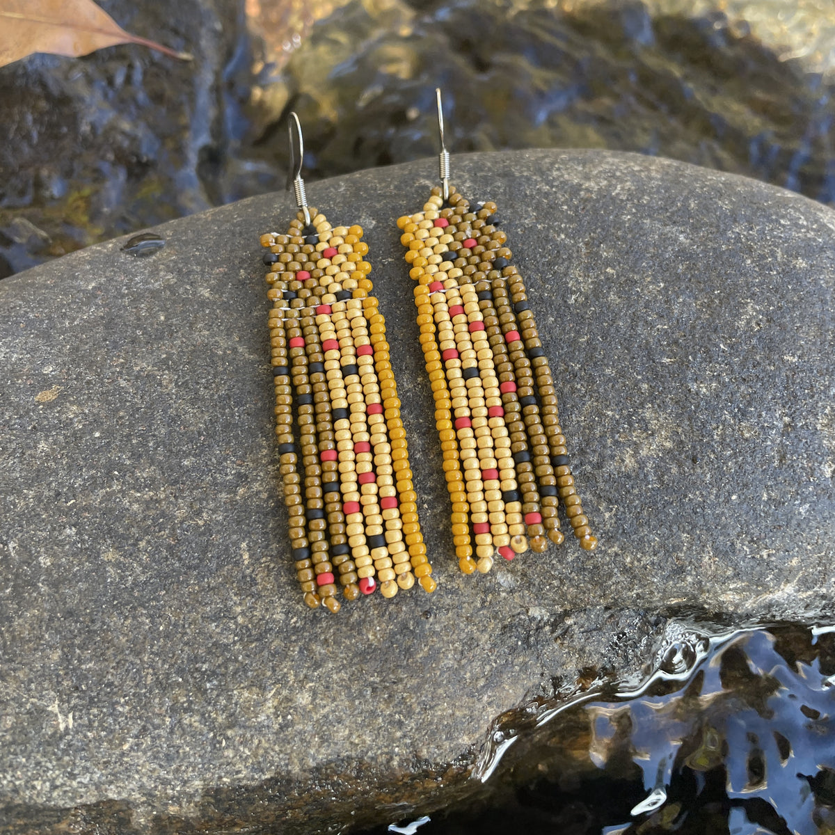 Pair of handmade beaded earrings in gold, red, and brown tones displayed on a rock next to flowing creek water.